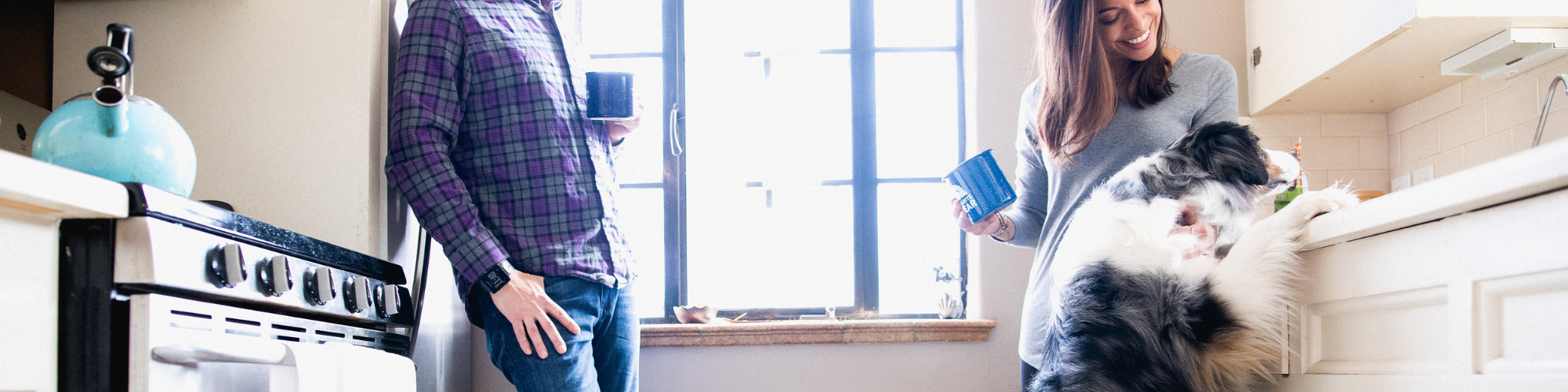 A man and woman stand in a bright kitchen with coffee mugs. The man leans against a stove, while the woman smiles at a large, fluffy dog with its front paws up on the counter. Sunlight streams through the window behind them.
