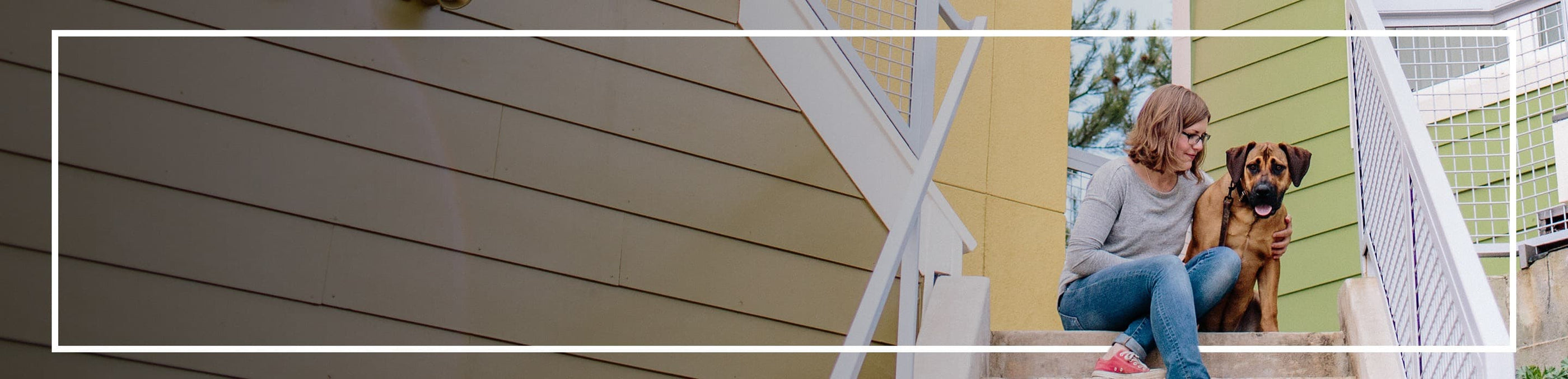 A blonde woman wearing glasses and a grey sweater sits on outdoor steps, affectionately placing her arm around a large brown dog. The background features colorful, modern house walls and railings.