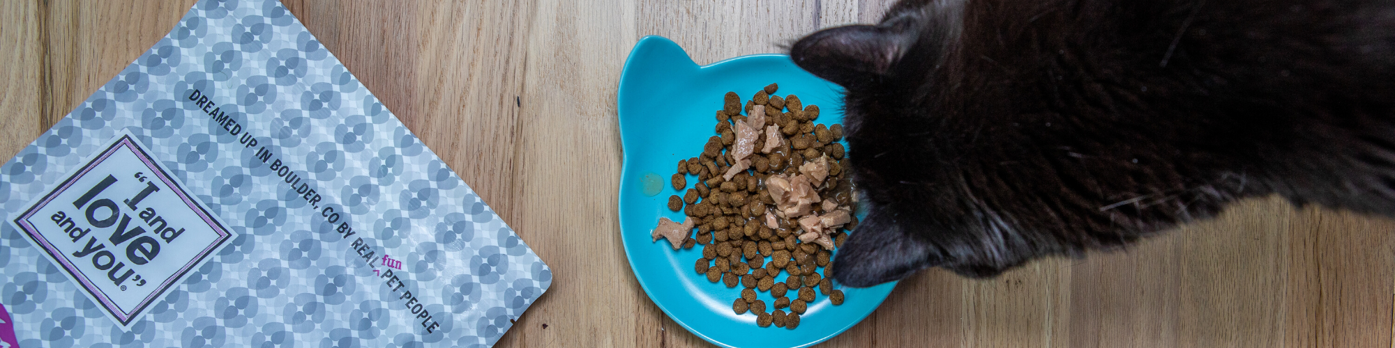 A black cat eats from a blue cat-shaped bowl filled with dry cat food and tuna flakes. The bowl sits on a wooden floor next to a gray and white patterned bag of dry cat food labeled “I and love and you.”.