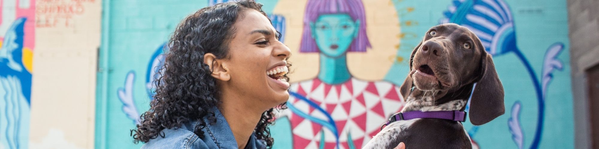 A woman laughing in front of a mural with a surprised dog nearby.