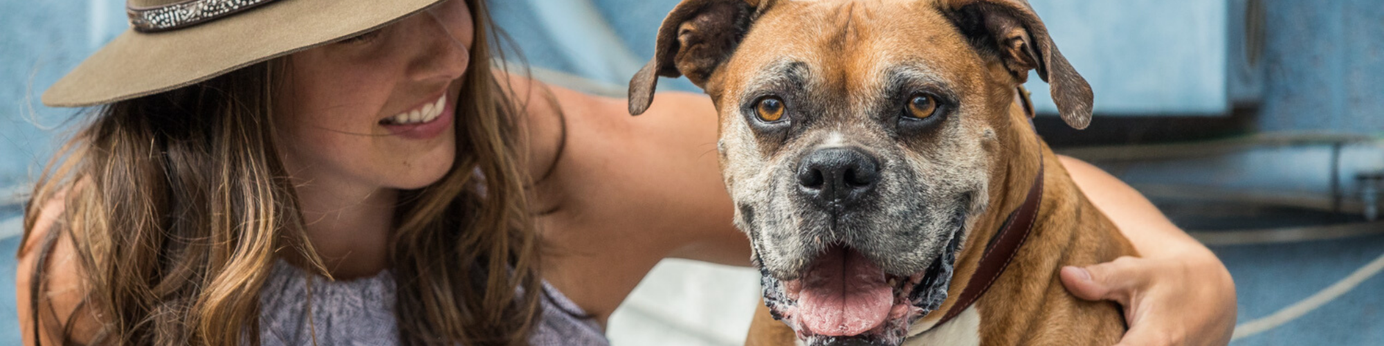 A woman wearing a hat and a light sleeveless top is smiling and looking at a brown and white Boxer dog. The dog, with a wide open mouth and visible teeth, also appears joyful. They are outdoors with a blurred background.