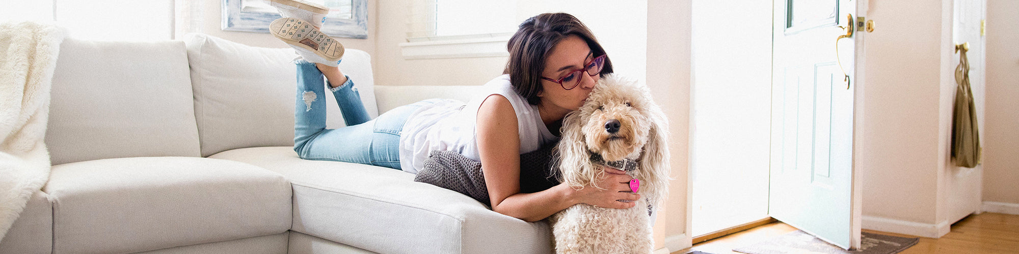 A woman with glasses is lying on a white sofa, propped up on one elbow, and hugging a fluffy dog. She is wearing a white shirt, ripped jeans, and sneakers, with one leg bent. The room is bright with a light-colored door slightly open in the background.