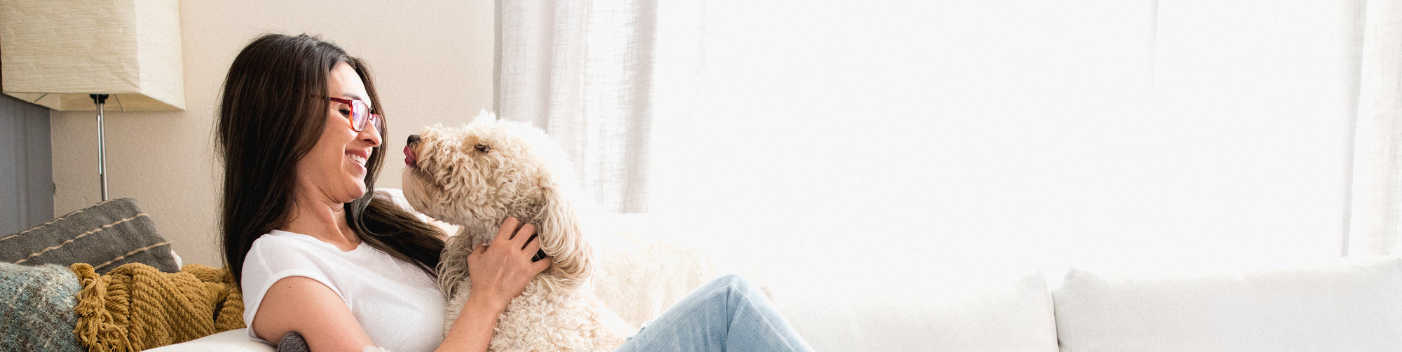 A woman with glasses and long hair is sitting on a white couch, smiling and holding a small, fluffy dog close to her face. The room is softly lit with natural light coming from a window in the background.