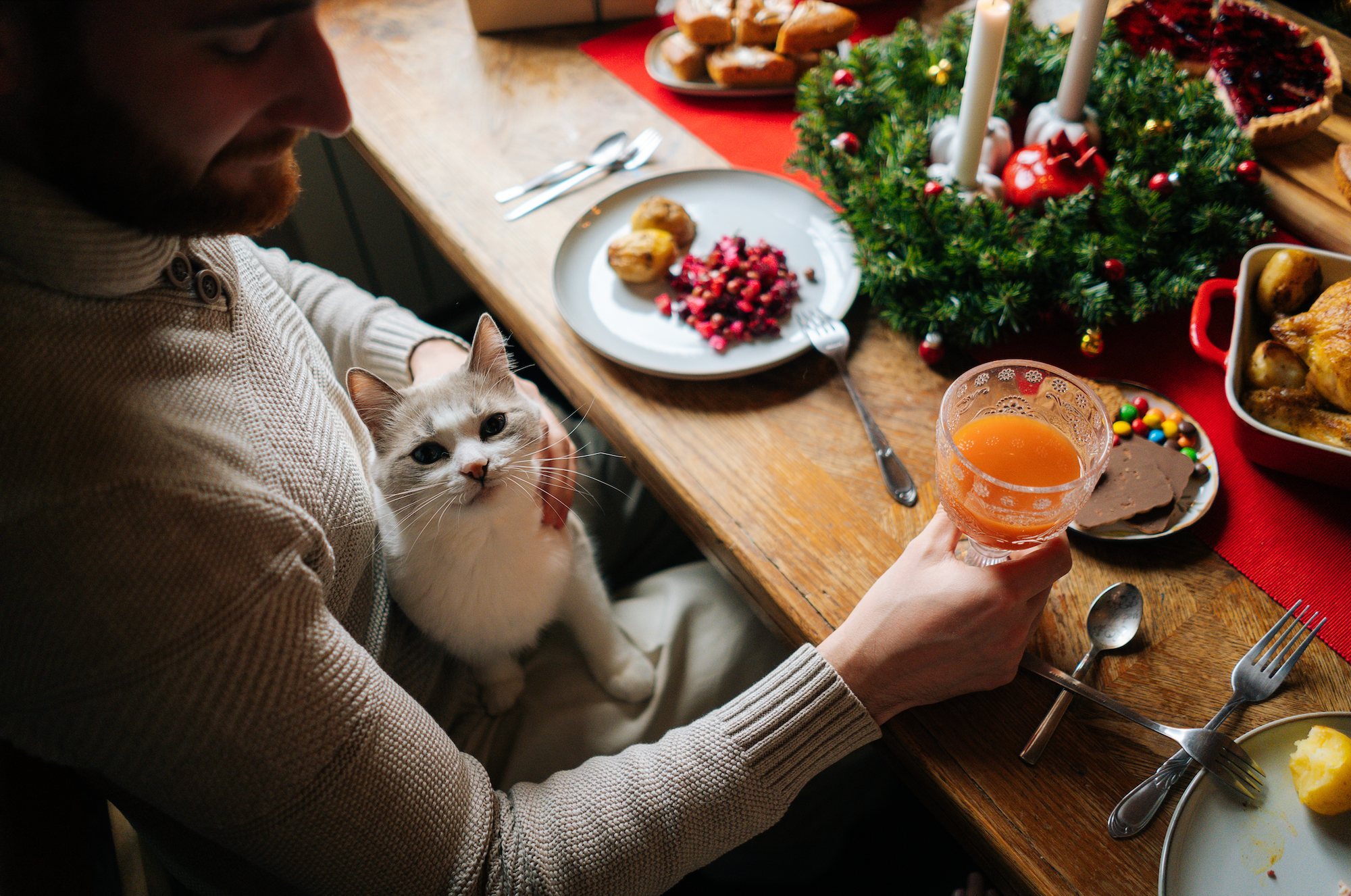 A man holding a glass of juice, petting a cat at a table.