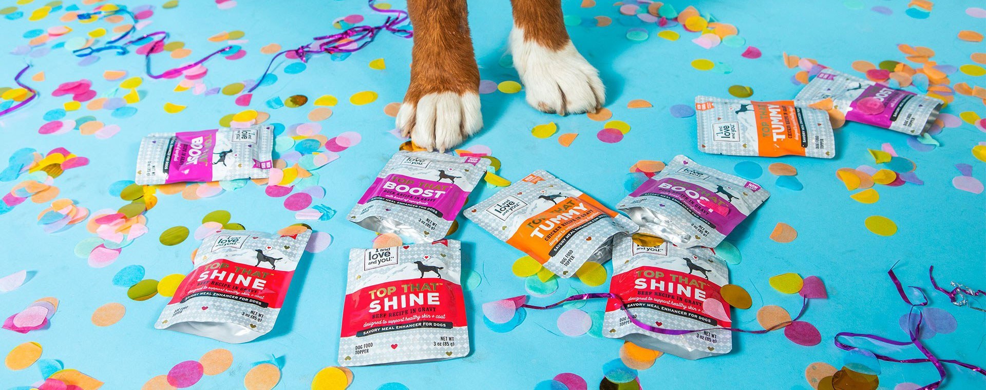 A dog's paws are surrounded by various packets of dog food toppers on a confetti-strewn blue surface. The packets come in different colors and flavors, including "Top That Shine," "Boost," and "Yummy." Colorful confetti and streamers are scattered around.