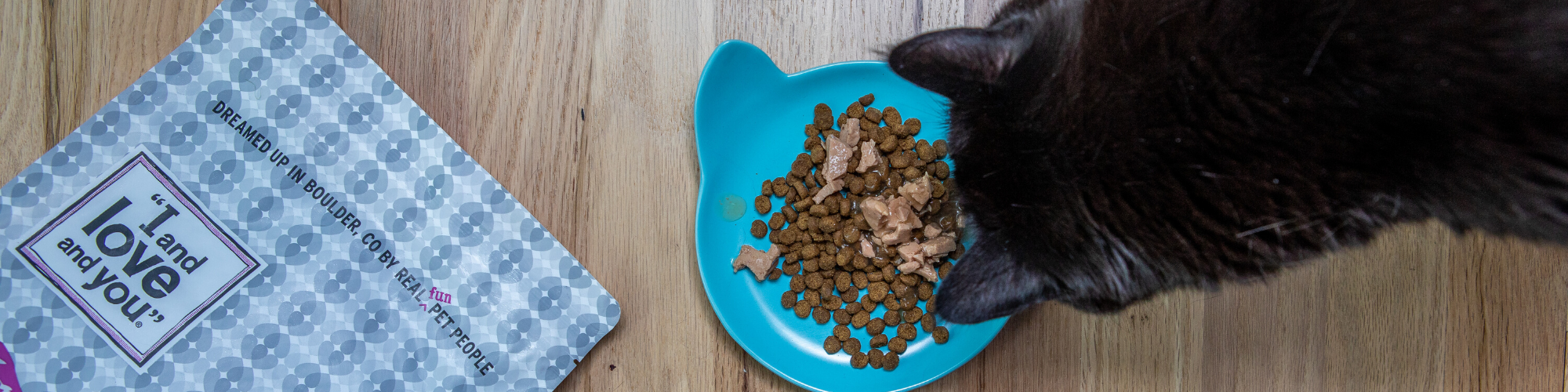 A black cat eats from a blue cat-shaped bowl filled with dry cat food and tuna flakes. The bowl sits on a wooden floor next to a gray and white patterned bag of dry cat food labeled ‚ÄúI and love and you.‚Äù.