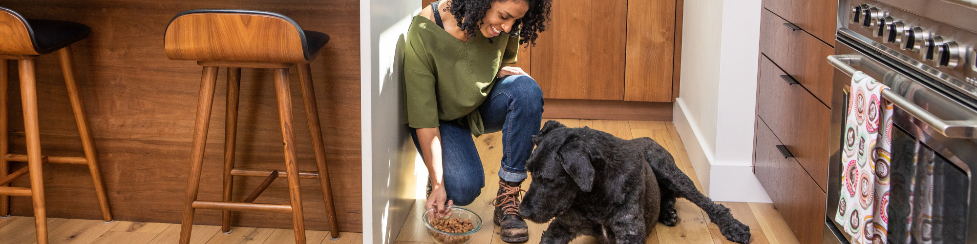 A person with curly hair is kneeling on a wooden floor in a kitchen, smiling and holding a bowl of dog food in front of a large black dog lying down. The kitchen features wooden cabinets and a stove with colorful towels hanging from the handle.