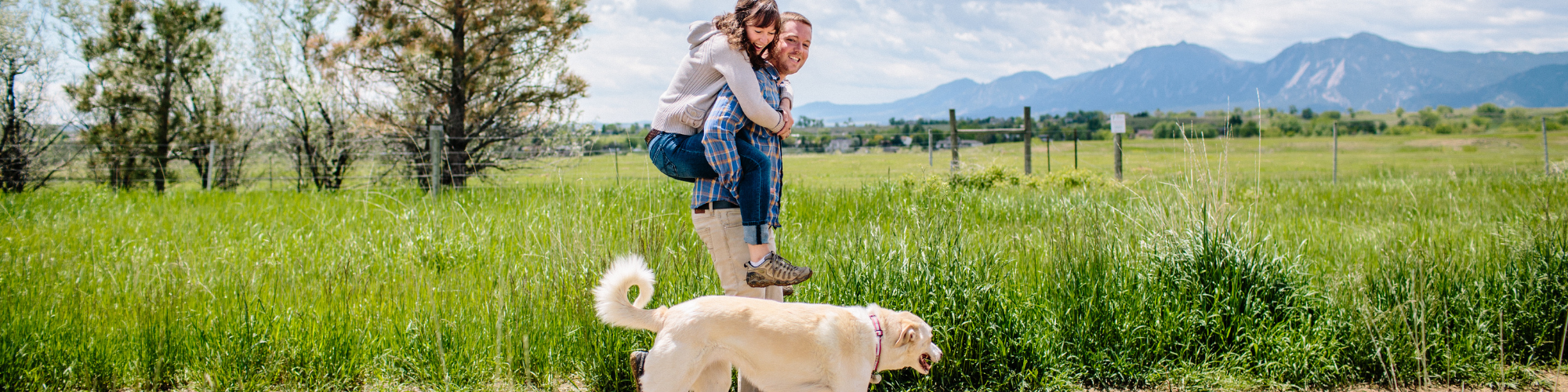 A person is giving a piggyback ride to another in a field with mountains in the background. They are both smiling and appear happy. A large, light-colored dog is walking in front of them. The field is lush and green, and the sky is partly cloudy.