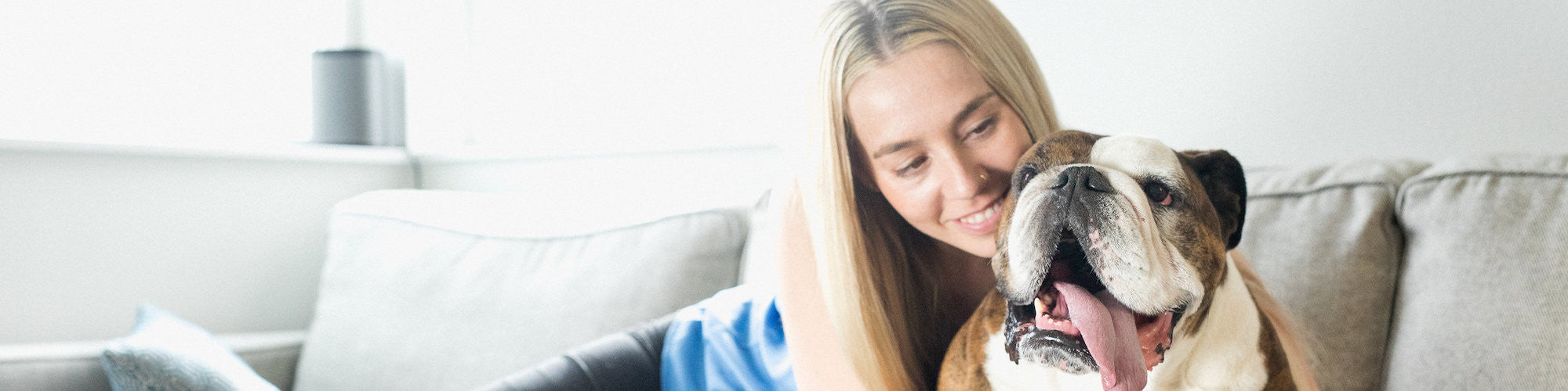 A person with long blonde hair is smiling and leaning on a couch next to a cheerful, panting bulldog. The person appears happy and relaxed. The background features a window and a lamp, creating a cozy indoor setting.