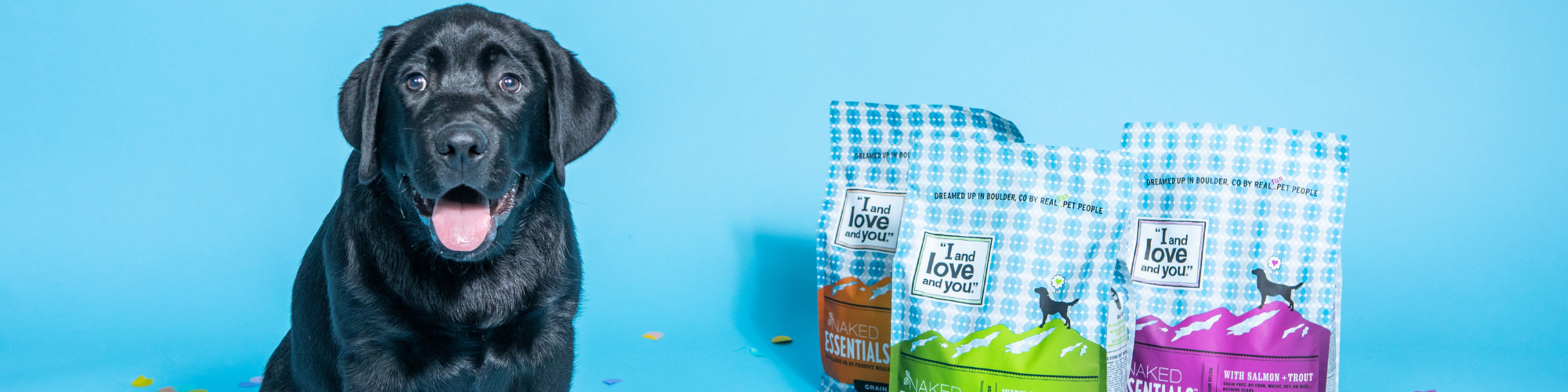 A happy black dog sits in front of three bags of 1 and Love and You brand dog food against a light blue background. The dog food bags are in different colors, featuring a mix of vegetables and meat ingredients illustrations.